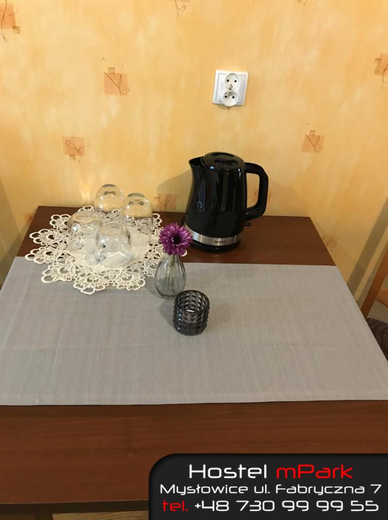 Stół w pokoju w Katowicach nakryty szarą bieżnikiem, na którym stoją szklanki, czajnik elektryczny, mały wazonik z kwiatkiem i świecznik. Na ścianie widoczne gniazdko elektryczne i wzorzysta tapeta.