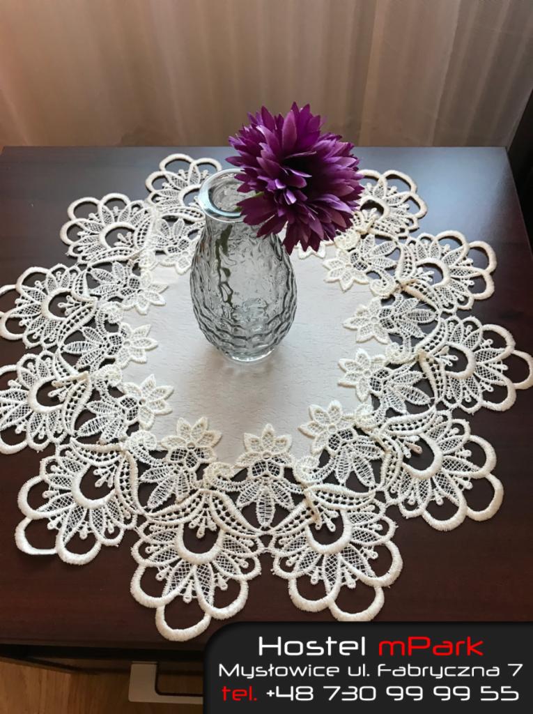 Dekoracyjny, okrągły bieżnik z koronki w kolorze ecru na ciemnym stole, z wazonem z fioletowym kwiatem w środku.