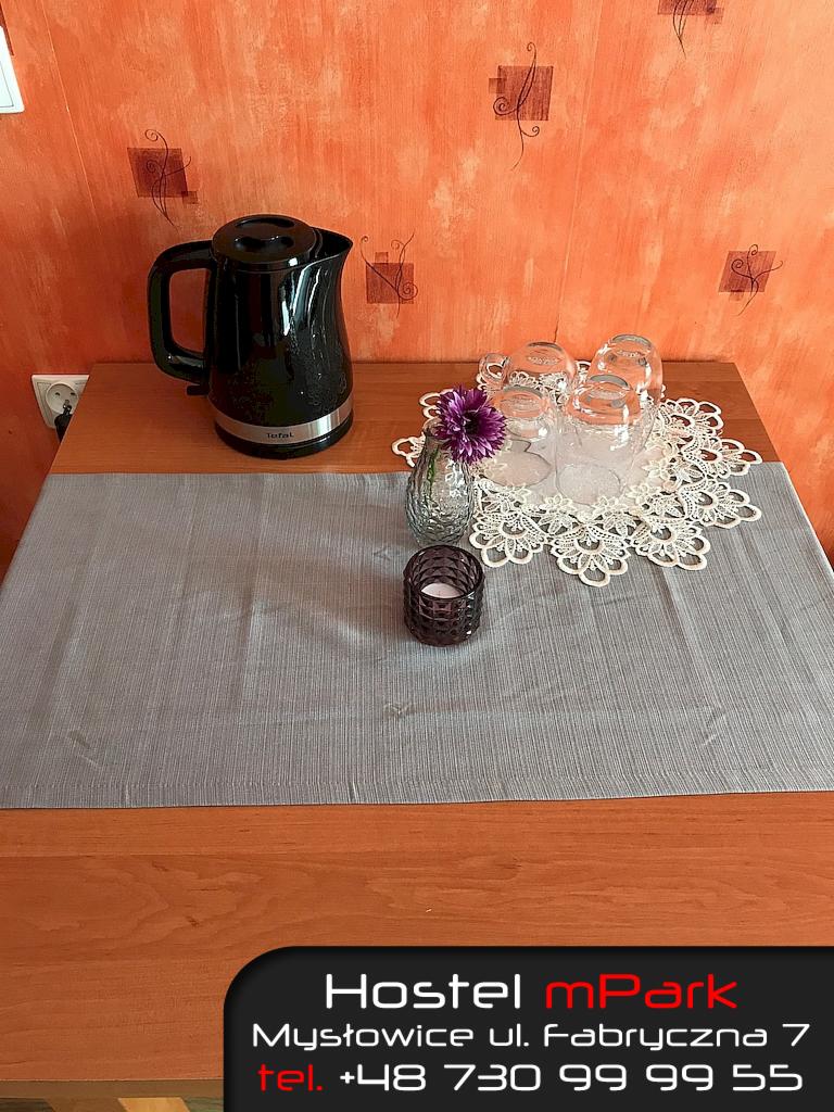 Stół w hostelu z czajnikiem elektrycznym, czterema szklankami na koronkowej serwetce, wazonikiem z fioletowym kwiatem i świecznikiem, na tle pomarańczowej ściany z dekoracjami.
