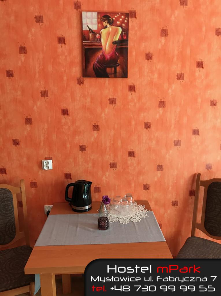 Pokój w hostelu z pomarańczowymi ścianami, stolikiem z czajnikiem i szklankami, nad którym wisi obraz z kobietą przy barze.