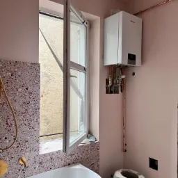 Łazienka w trakcie remontu: biała wanna, ściany w różowym odcieniu z terazzo, zamontowany piec gazowy Vaillant z miedzianymi rurami, otwarte okno z widokiem na zewnątrz, zakurzony taboret.