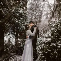 Zakochana para całuje się w zimowym lesie podczas sesji narzeczeńskiej, na tle ośnieżonych drzew i padającego śniegu.