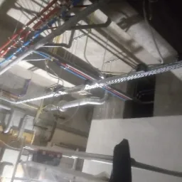 Sufit w trakcie montażu instalacji: widoczne rury wodne w kolorach czerwonym i niebieskim, kable elektryczne w korytkach oraz fragment drabiny z narzędziami.