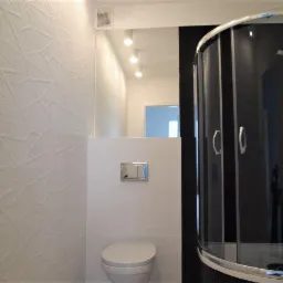 Nowoczesna łazienka z białą mozaikową ścianą, podwieszaną toaletą i kabiną prysznicową z czarnymi elementami.