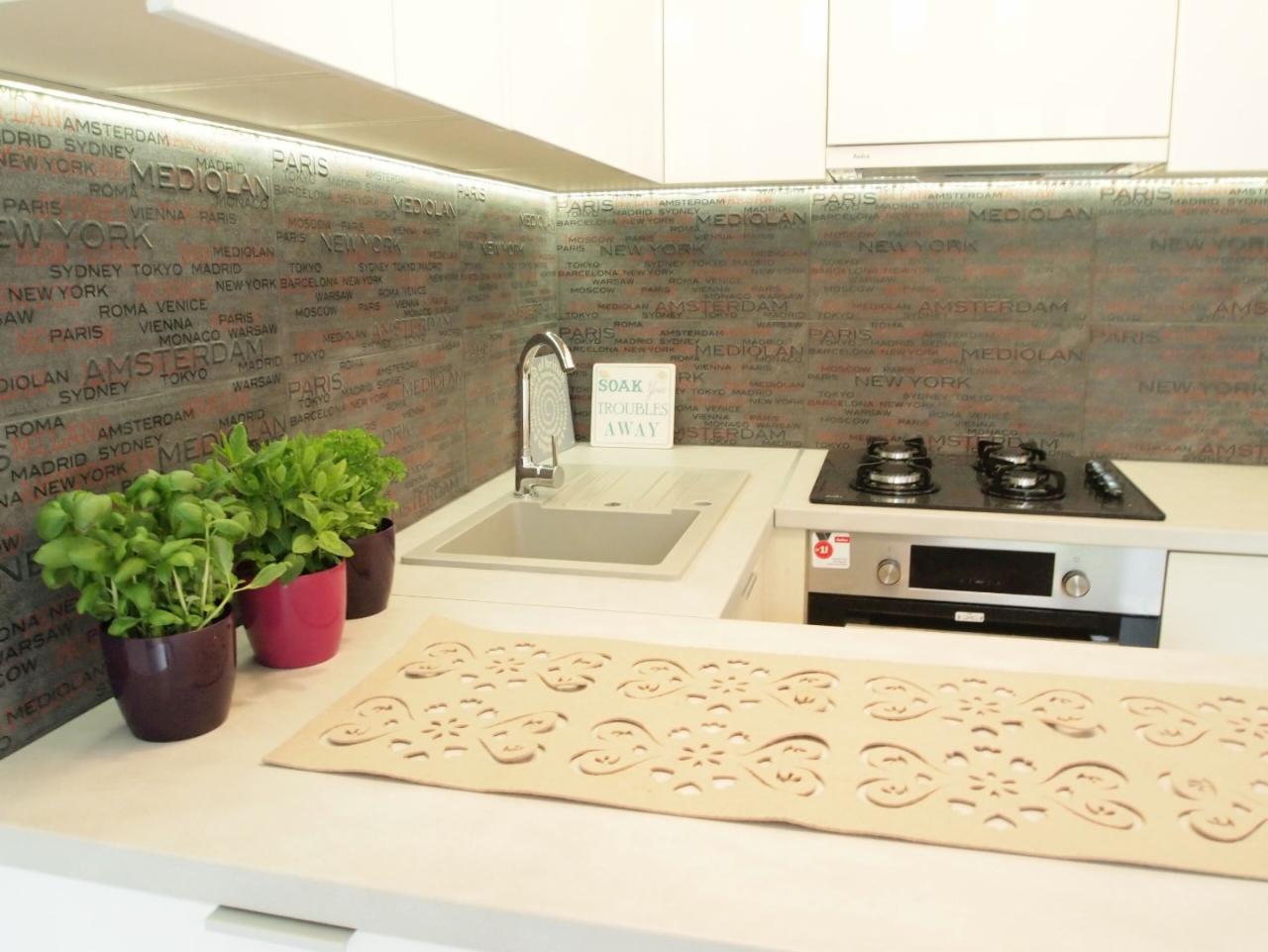 Nowoczesna kuchnia z białymi szafkami, szarym blatem i płytkami z motywem mapy miast na ścianie. Na blacie stoją doniczki z zielonymi roślinami i dekoracyjna serwetka z wycinanym wzorem.