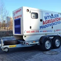 Biały agregat prądotwórczy na przyczepie, z niebieskimi akcentami i napisem 'Wynajem Agregatów Prądotwórczych', zaparkowany na brukowanym placu w słoneczny dzień.