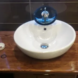 Biała, okrągła umywalka nablatowa z chromowaną baterią na blacie z drewna sosnowego o wyraźnym usłojeniu, obok leżą biała osłonka i czarny oring, tło stanowią płytki i tapeta w kwiatowy wzór.