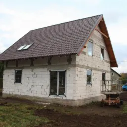 Niedokończony dwukondygnacyjny budynek z białego bloczka z brązowym dachem i oknami PCV, widoczny podnośnik koszowy, w tle zabudowa jednorodzinna i pochmurne niebo.