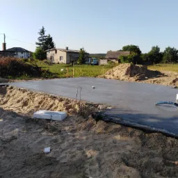 Świeżo wylana, gładka płyta betonowa fundamentowa otoczona piaskiem i folią izolacyjną, z widocznymi elementami instalacji i rurami, na tle wiejskiego krajobrazu z domami.