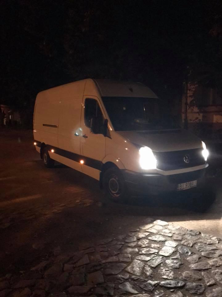 Biały bus Mercedes Sprinter z włączonymi światłami na brukowanej drodze nocą. Widoczna tablica rejestracyjna.