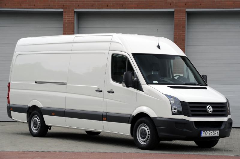 Biały Volkswagen Crafter, widok z przodu i boku, zaparkowany na kostce brukowej przed budynkiem z cegły i szarymi bramami garażowymi.