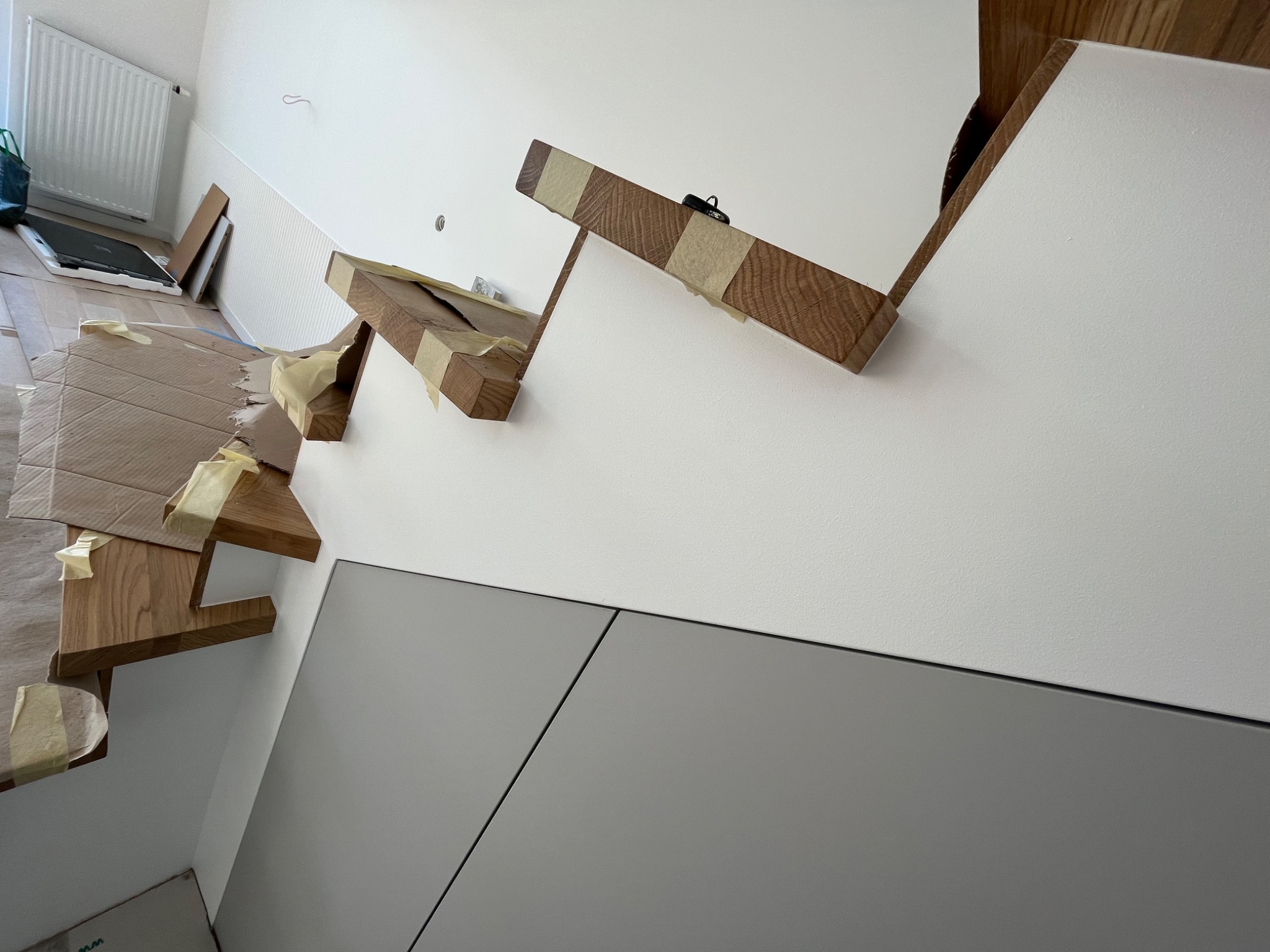 Montaż drewnianych schodów z użyciem taśmy malarskiej, widoczne szare panele ścienne i biała ściana z fakturą.