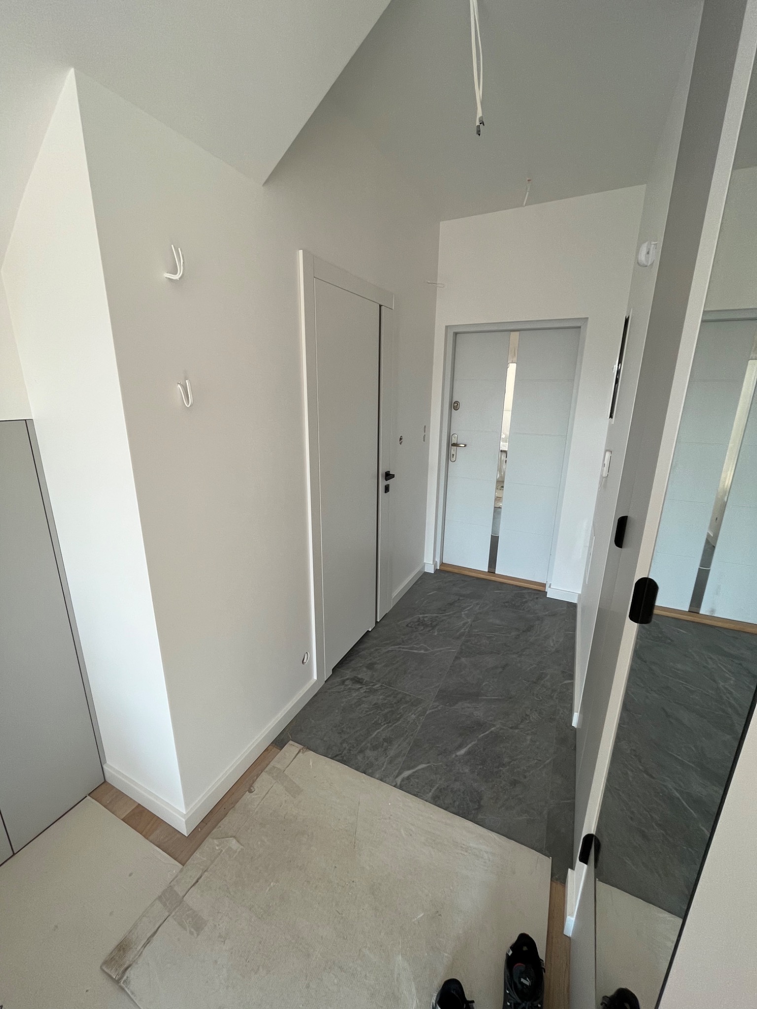 Jasne wnętrze przedpokoju w trakcie prac wykończeniowych, widoczne szare płytki podłogowe, białe ściany z wieszakami i dwoje białych drzwi, podłoga zabezpieczona kartonem.