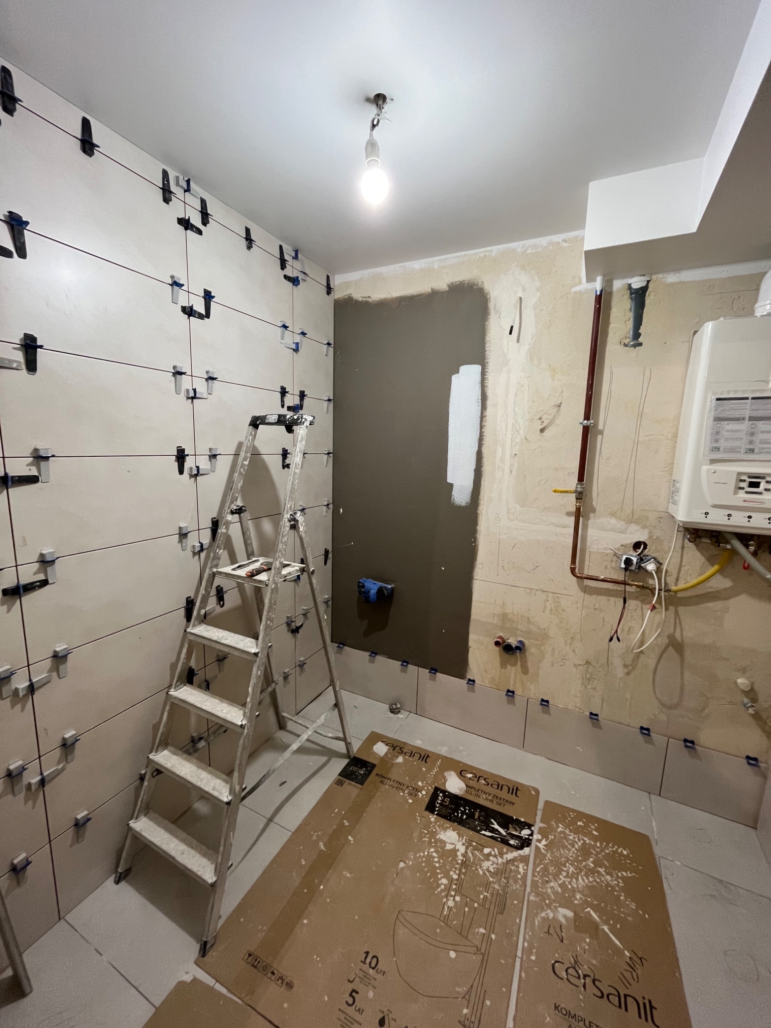 Łazienka w trakcie układania płytek, widoczne listwy wyrównujące, drabina aluminiowa, kartony po ceramice Cersanit na podłodze, fragment ściany przygotowany pod malowanie, instalacja grzewcza.