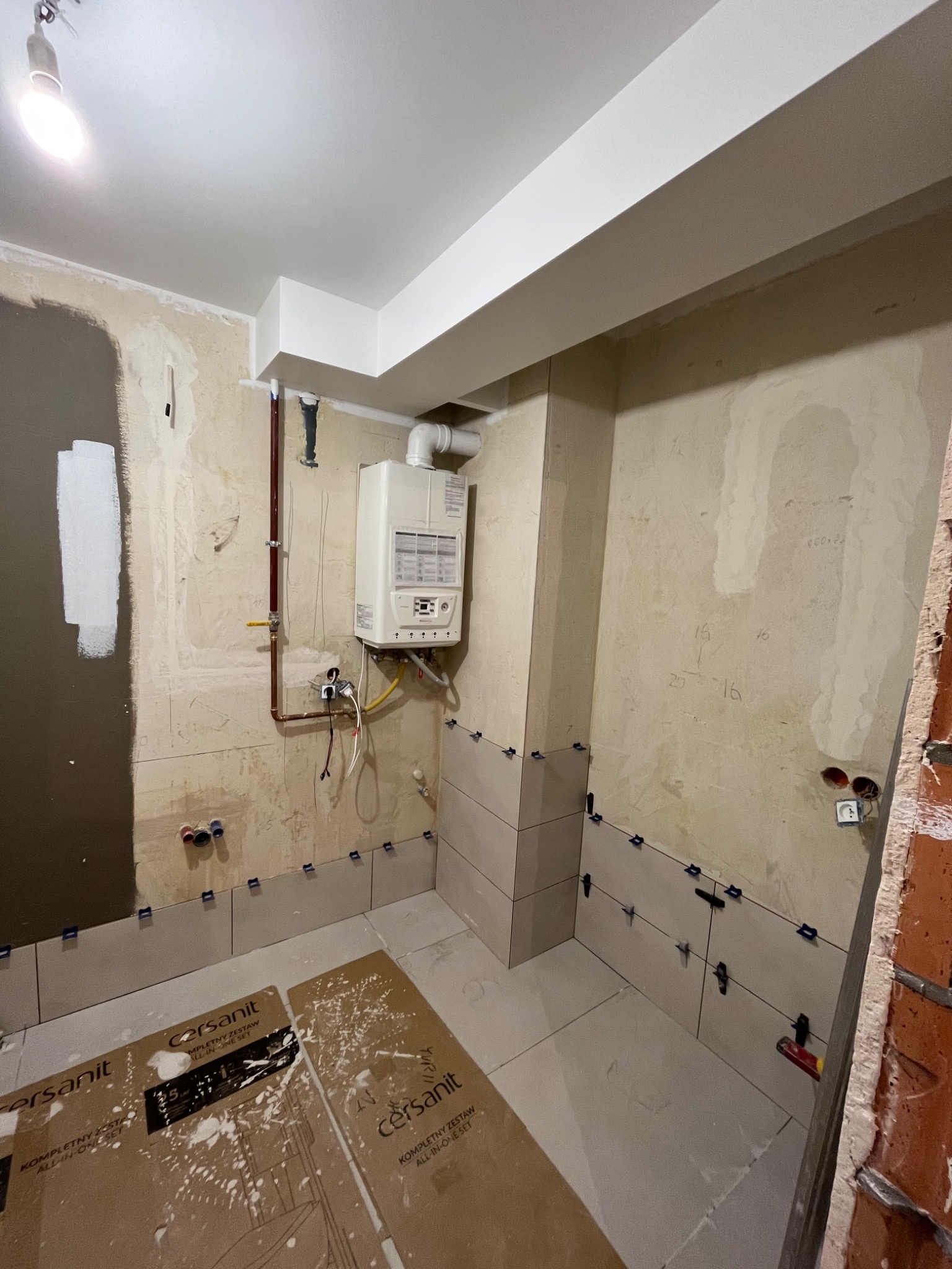 Remont łazienki w trakcie: częściowo ułożone jasne płytki na ścianach i podłodze, widoczny piec gazowy, kartony po zestawie Cersanit, nieotynkowane ściany z zaznaczeniami, surowa cegła w rogu