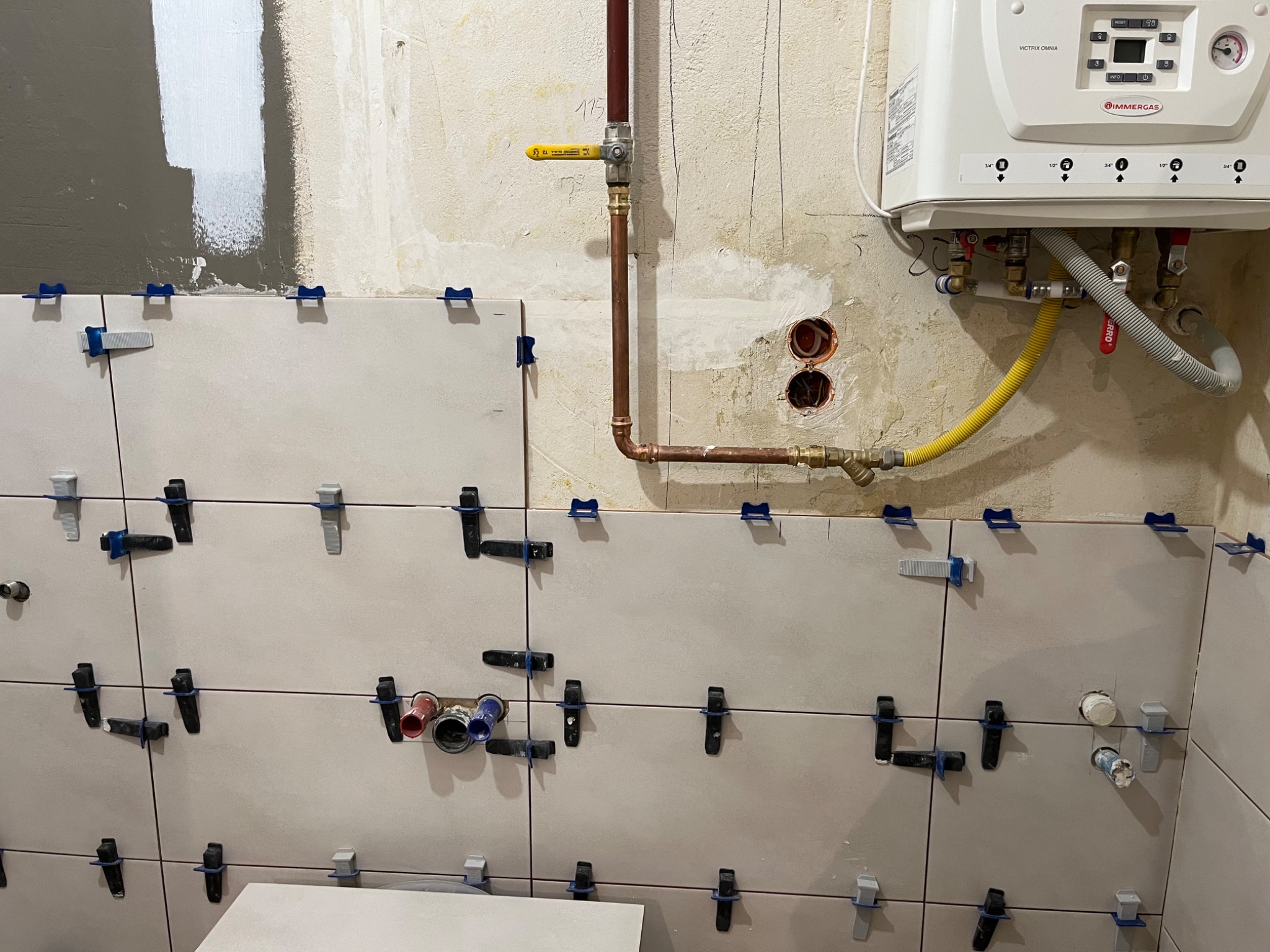 Ściana łazienki w trakcie układania płytek, widoczne krzyżyki dystansowe, fragment instalacji wodnej i piec gazowy Immergas na ścianie w tle.