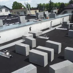 Montaż systemu mocowania paneli na dachu płaskim z użyciem betonowych obciążników, widoczny pracownik w oddali, drabina oparta o budynek.