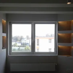 Wykończony pokój z gładzią na ścianie o szarej, nieregularnej fakturze, podświetlane półki wnękowe, okno z widokiem na budynki i drzewa, biały sufit z dekoracyjnymi belkami i oświetleniem punktowym.