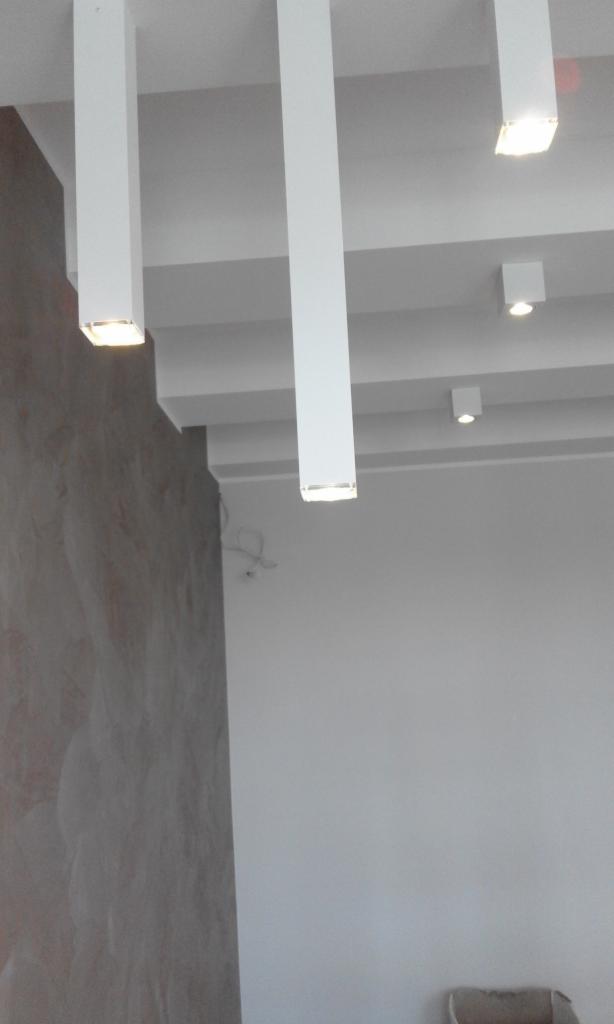 Nowoczesny sufit z podwieszanymi, prostokątnymi lampami LED i minimalistycznymi punktami świetlnymi, ściana z dekoracyjną fakturą.