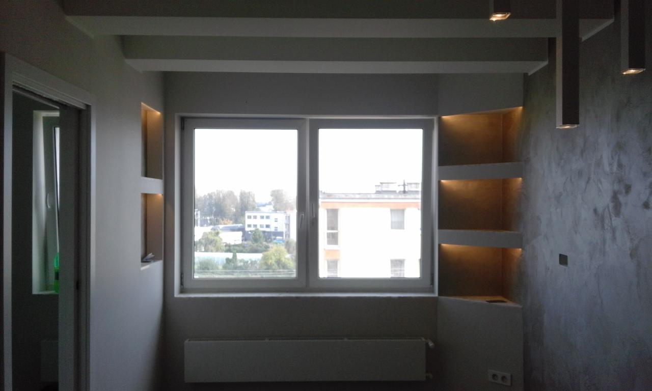 Wykończony pokój z gładzią na ścianie o szarej, nieregularnej fakturze, podświetlane półki wnękowe, okno z widokiem na budynki i drzewa, biały sufit z dekoracyjnymi belkami i oświetleniem punktowym.