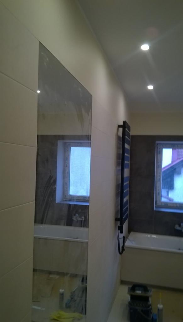 Wnętrze łazienki w trakcie prac wykończeniowych, widoczna ściana z jasnymi płytkami, fragment lustra, grzejnik drabinkowy, okno oraz akcesoria budowlane na podłodze.