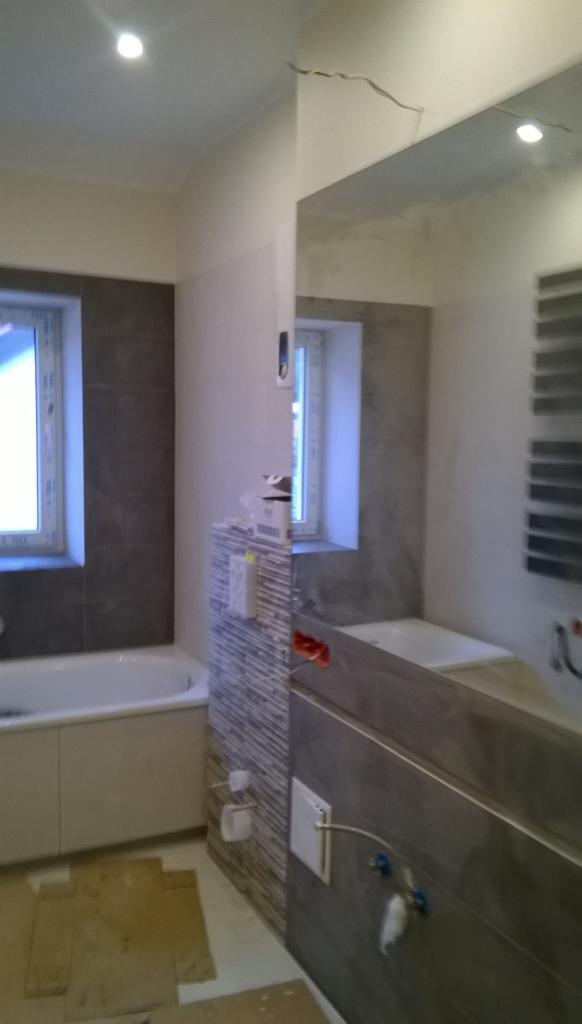 Łazienka w trakcie remontu: częściowo wykończone ściany z szarymi płytkami, widoczne instalacje hydrauliczne i elektryczne, zabezpieczona podłoga, lustro odbija fragment łazienki.