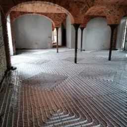 Instalacja ogrzewania podłogowego w piwnicy z ceglanym sklepieniem łukowym, rury grzewcze ułożone na folii izolacyjnej, widoczne kolumny podtrzymujące sklepienie.