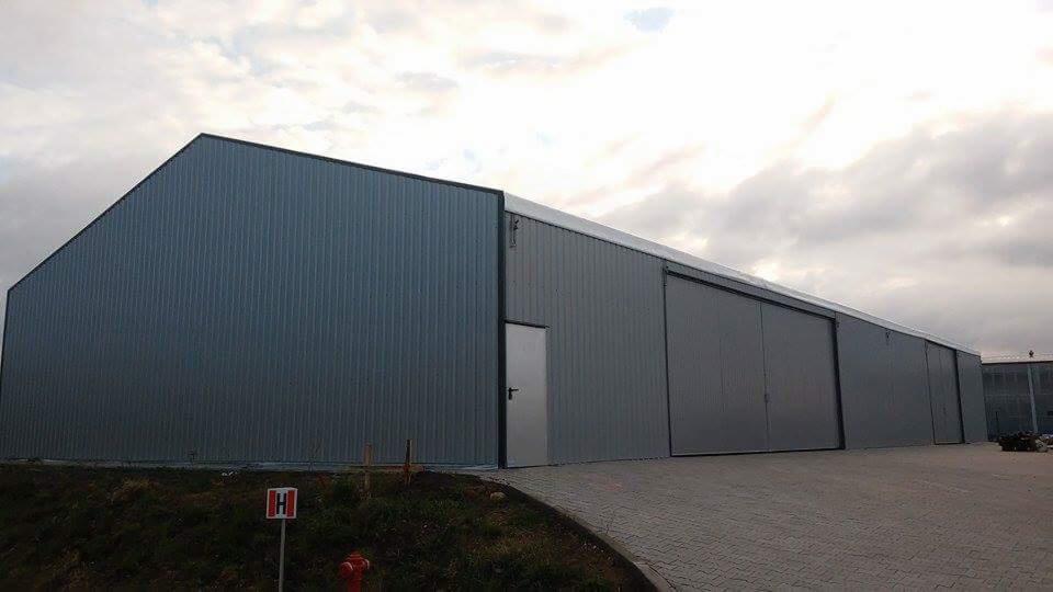 Gotowa hala magazynowa z szarej blachy trapezowej z przesuwanymi drzwiami i małą furtką wejściową, widoczny znak hydrantu przeciwpożarowego na pierwszym planie.