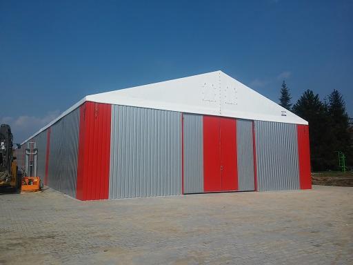 Gotowa hala magazynowa z płyty warstwowej, pomalowana w pasy czerwono-szare, z białym dachem, na zewnątrz stoi pomarańczowy wózek widłowy.