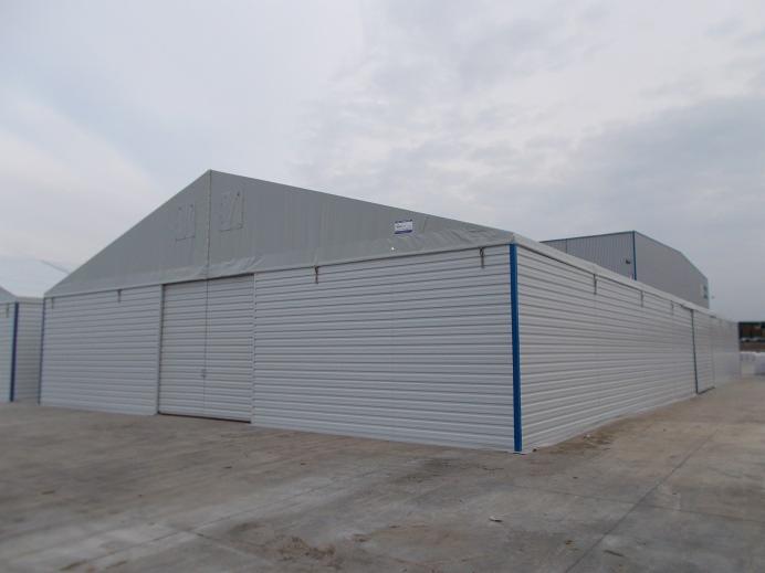 Szeroki, niski magazyn z płyt warstwowych, szare ściany z poziomymi przetłoczeniami, błękitne obramowanie dolnej krawędzi, przesuwne drzwi na środku, pochmurne niebo w tle.