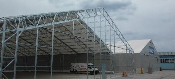 Szkielet konstrukcji stalowej hali w trakcie budowy, widoczny biały samochód dostawczy z logo firmy na tle szarej, gotowej części hali.