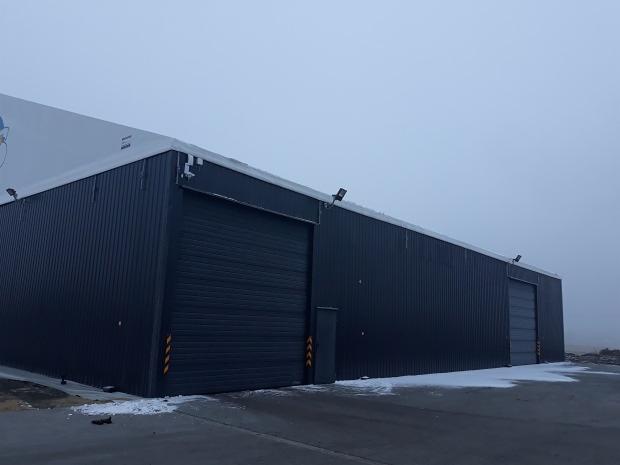 Gotowa hala przemysłowa z płyt warstwowych w pochmurny dzień, widoczne dwie bramy segmentowe i oświetlenie zewnętrzne.