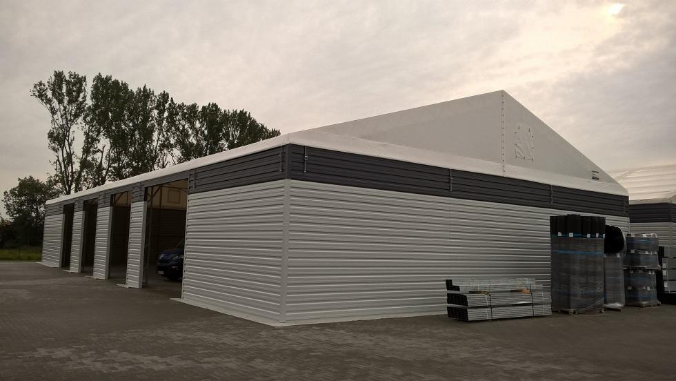 Gotowa hala magazynowa z szarej blachy trapezowej z segmentowymi bramami garażowymi, częściowo zadaszona białą plandeką, na zewnątrz ułożone materiały budowlane.