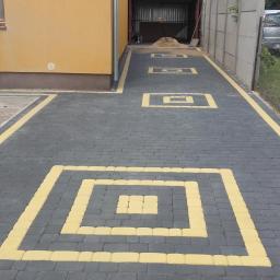 K-BRUK - Ułożona nawierzchnia z ciemnoszarej kostki brukowej z żółtymi akcentami w kształcie kwadratów, prowadząca do garażu, widoczne dwa samochody w tle.