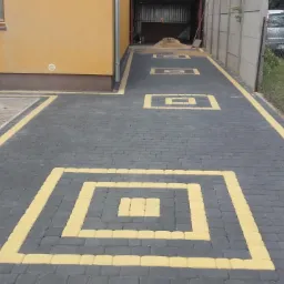 Ułożona nawierzchnia z ciemnoszarej kostki brukowej z żółtymi akcentami w kształcie kwadratów, prowadząca do garażu, widoczne dwa samochody w tle.