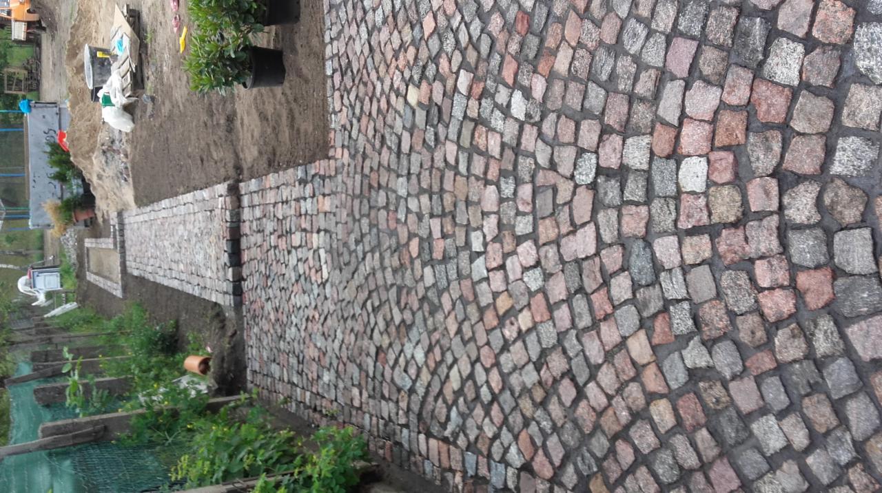 Ścieżka z kostki granitowej ułożona w łukowaty wzór, otoczona ziemią i zielenią, widok z góry.