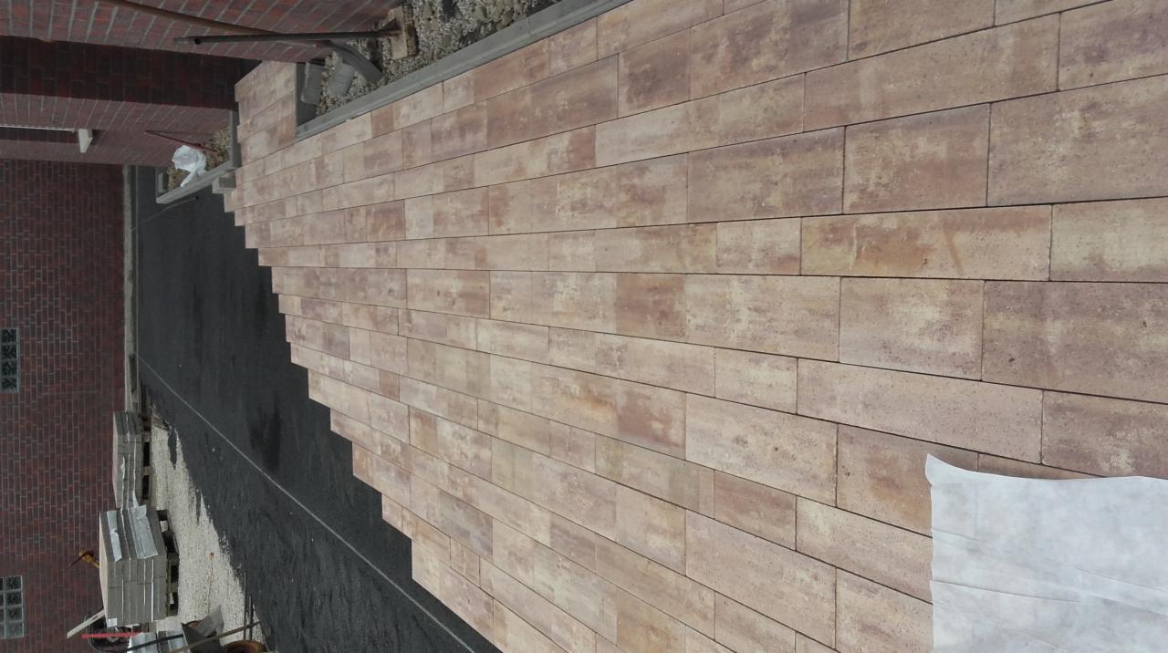 Widok z góry na układaną nawierzchnię z prostokątnej kostki w odcieniach beżu i brązu, widoczna czarna podsypka i fragment budynku z czerwonej cegły.