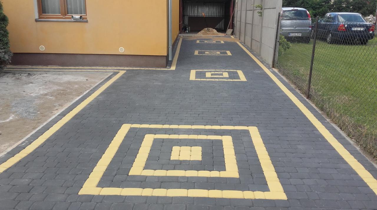 Ułożona nawierzchnia z ciemnoszarej kostki brukowej z żółtymi akcentami w kształcie kwadratów, prowadząca do garażu, widoczne dwa samochody w tle.