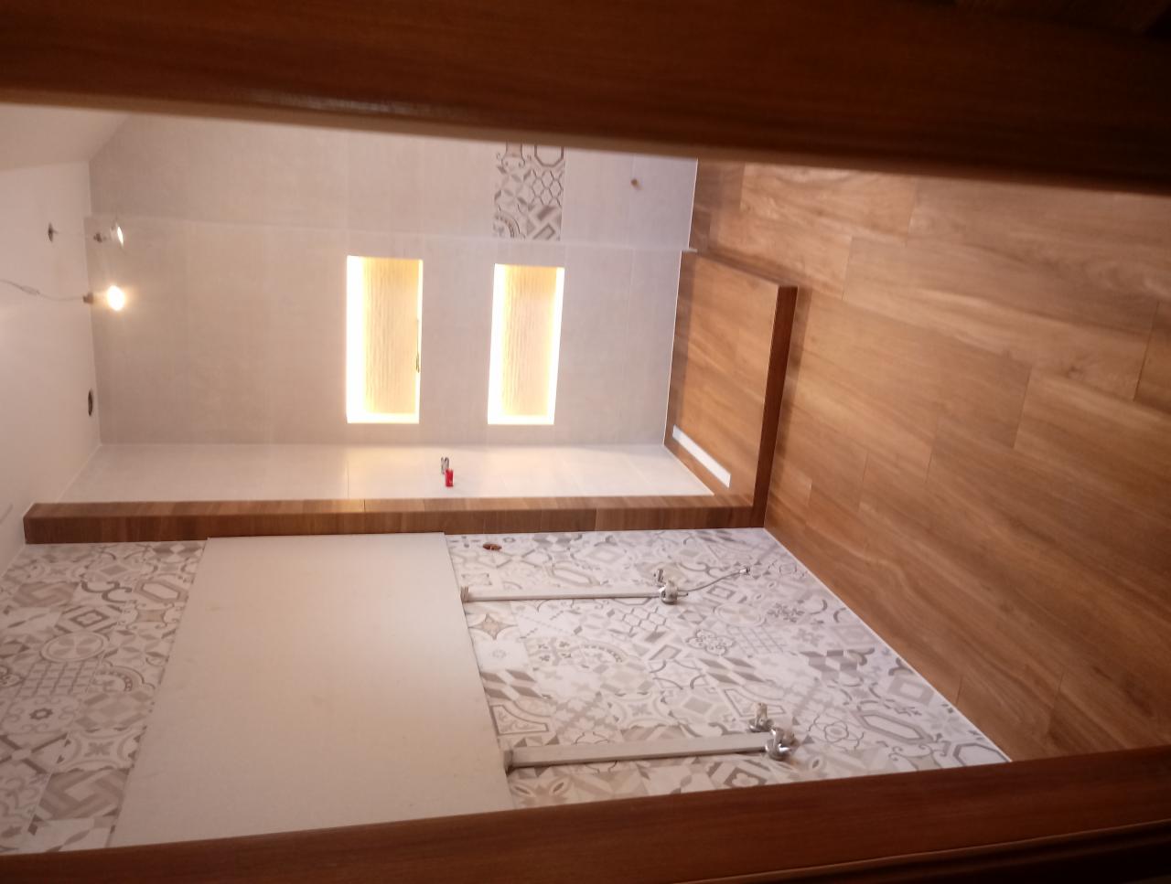 Wnętrze łazienki w trakcie wykańczania: częściowo ułożone dekoracyjne płytki podłogowe, drewnopodobna okładzina ścienna, widoczne profile montażowe i dwa podświetlane wnęki w ścianie.