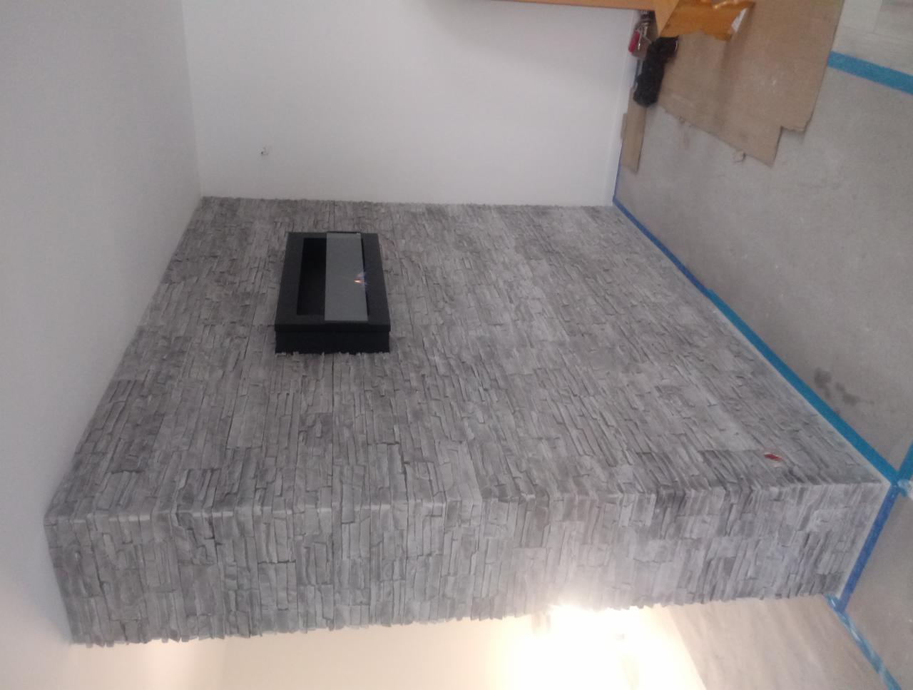 Ściana wykończona szarą okładziną imitującą kamień, z wbudowanym czarnym biokominkiem, widoczne zabezpieczenia taśmą malarską, fragment podłogi z kartonem ochronnym.