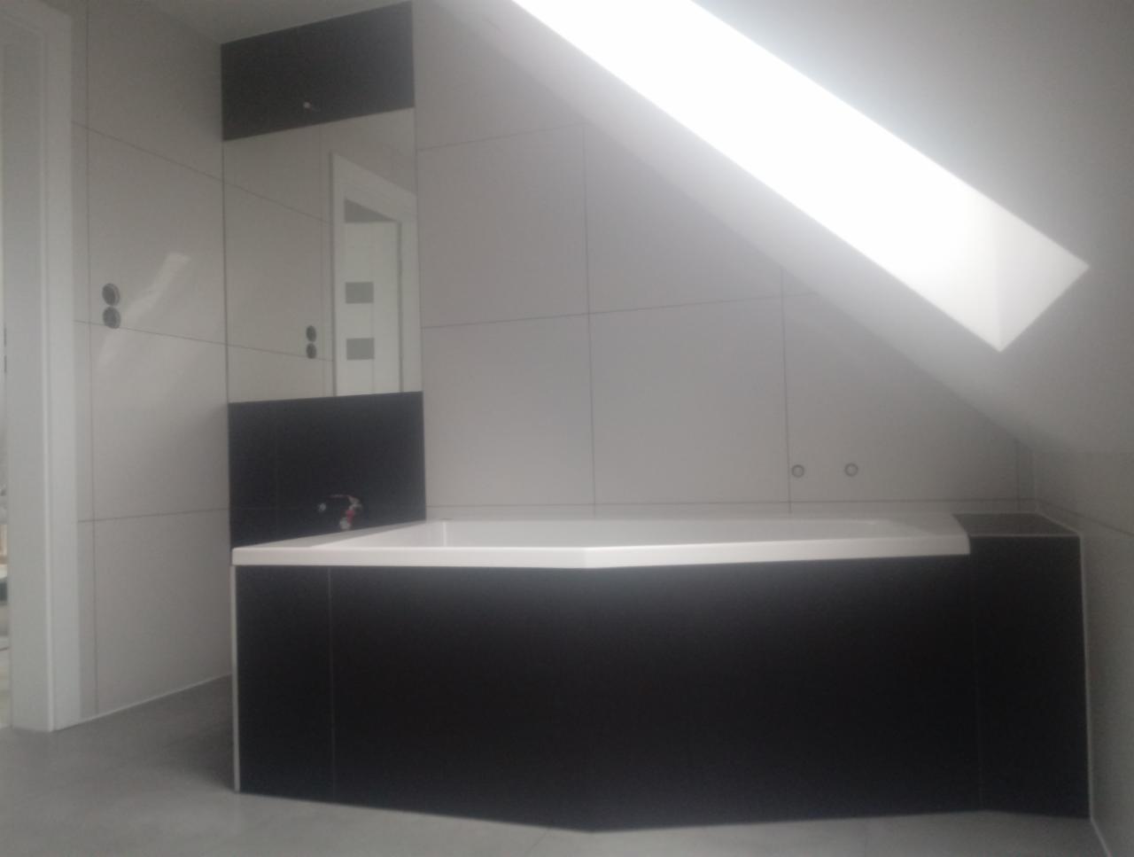 Wykończona łazienka na poddaszu z biało-czarnymi płytkami, obudowana wanna z czarną obudową, widoczne białe fugi i skosy dachu z oknem dachowym.