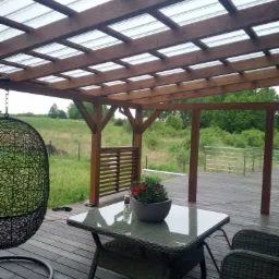 Zadaszony drewniany taras z meblami ogrodowymi, wiszącym fotelem i widokiem na zielony krajobraz w tle.