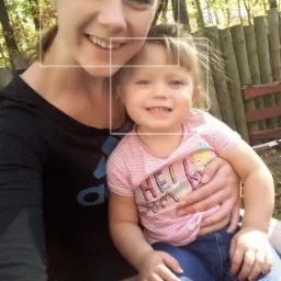 Uśmiechnięta młoda kobieta robi selfie z małą dziewczynką na kolanach, oboje patrzą w obiektyw na tle jesiennego krajobrazu.