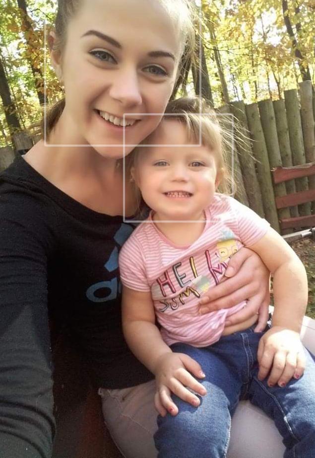 Uśmiechnięta młoda kobieta robi selfie z małą dziewczynką na kolanach, oboje patrzą w obiektyw na tle jesiennego krajobrazu.