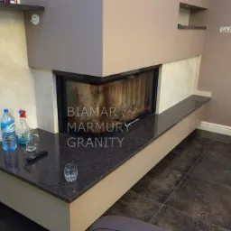 Nowoczesny kominek z obudową z ciemnego granitu, na którym stoją butelka wody, płyn do czyszczenia i pilot do telewizora, a w palenisku widać drewno.