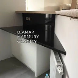 Błyszczący, czarny blat na białej szafce, z widocznym grzejnikiem i fragmentem ceglanej ściany w tle.