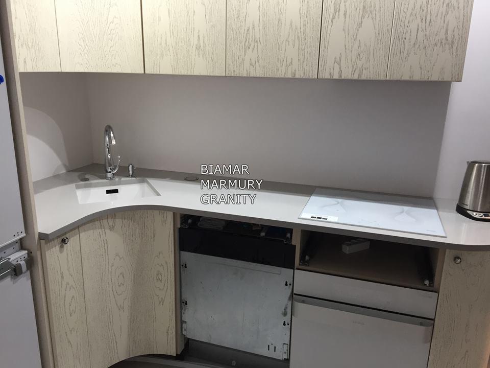 Jasny blat kamienny z wbudowanym zlewozmywakiem i płytą indukcyjną w nowoczesnej kuchni z drewnianymi szafkami, widoczny otwarty front szafki pod zlewem.