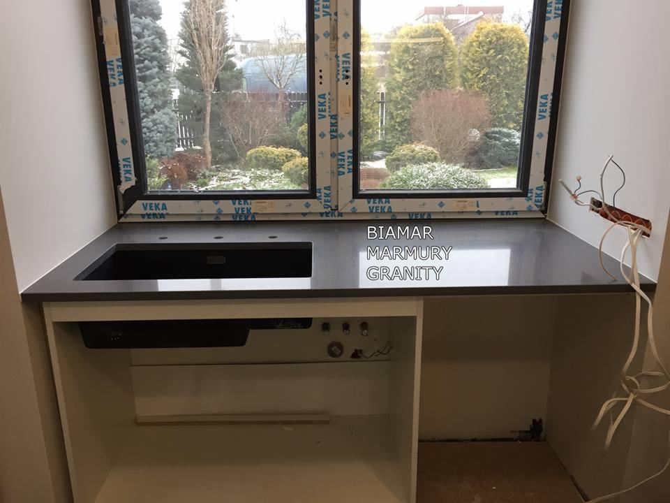 Ciemnoszary blat kuchenny z wbudowanym zlewozmywakiem, widok na okno z ogrodem zimowym, instalacja elektryczna w trakcie podłączania.
