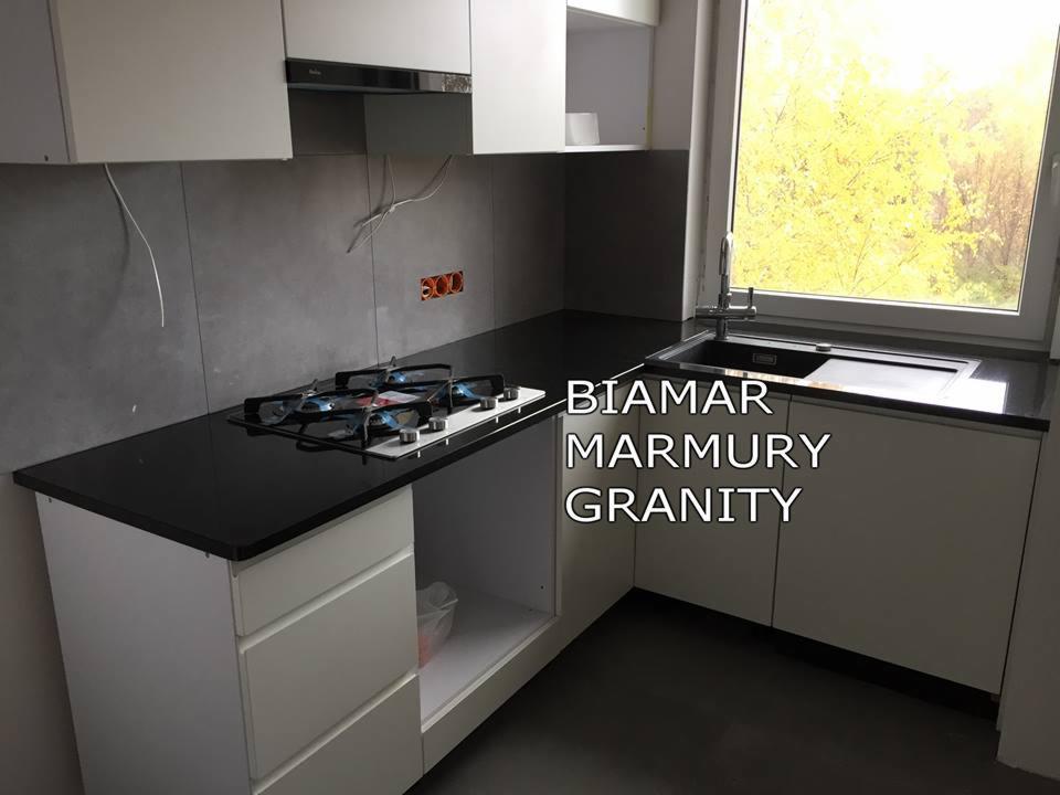 Czarny, błyszczący blat kuchenny z kamienia, zamontowany w nowoczesnej kuchni z białymi szafkami i widokiem na drzewa za oknem, z widoczną płytą gazową.