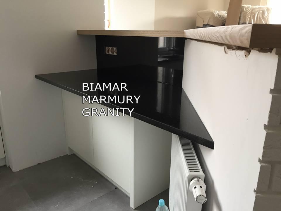 Błyszczący, czarny blat na białej szafce, z widocznym grzejnikiem i fragmentem ceglanej ściany w tle.